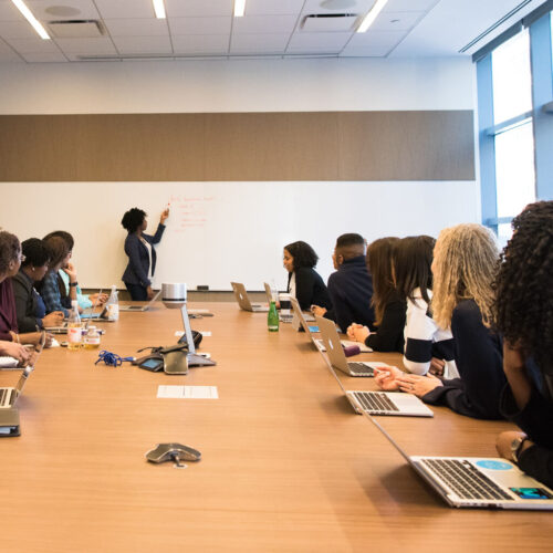 A photo of a conference room with various people sitting around the table with their laptops open and one person at the front of the room pointing to illegible content on a whiteboard